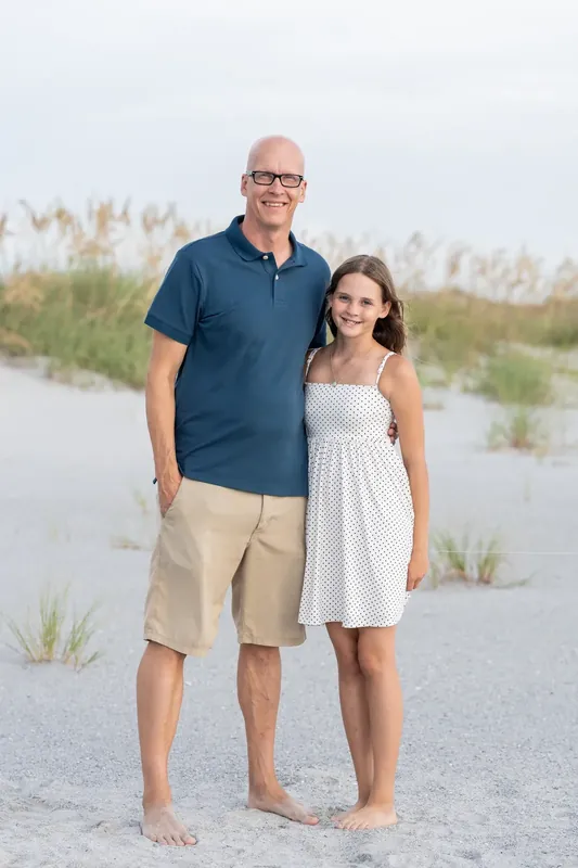 Man in a navy polo and khaki shorts stands beside a younger person in a white dress on the beach.
