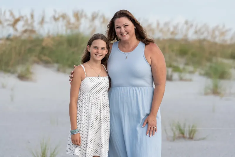 Woman in a light blue dress and a younger person in a white dress smile together with coastal dunes in the background.
