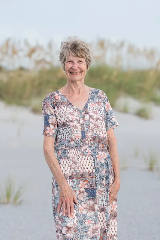 Kelly smiles at Vilano Beach with sea oats and dunes behind her, wearing a patterned floral dress.