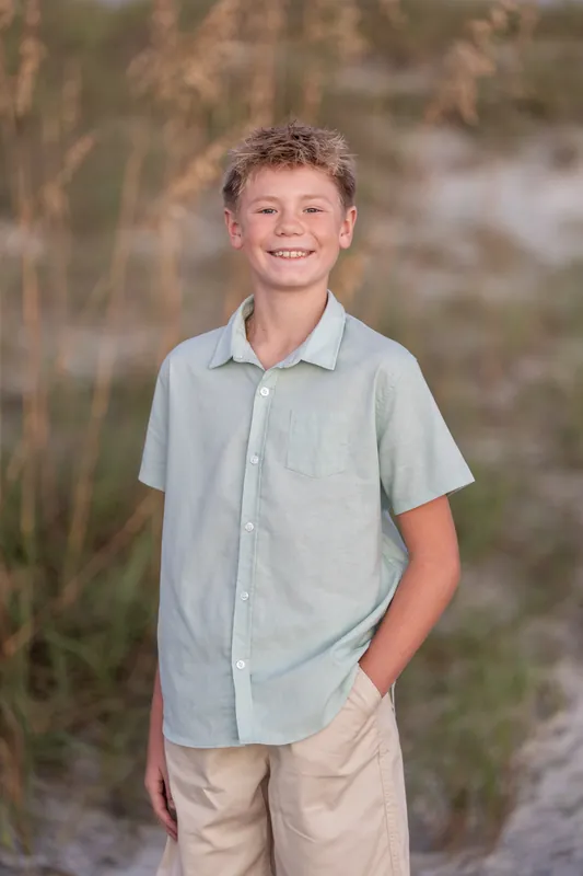 A young person in a light blue button-up shirt and khaki pants smiles at Vilano Beach with beach grasses behind.