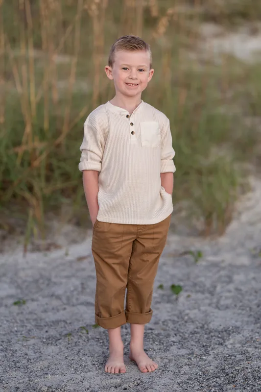 A child in a cream henley and brown cropped pants stands barefoot on the sand at Vilano Beach.