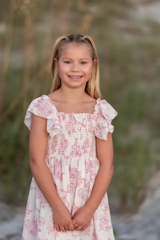A girl in a pink-and-white patterned dress with ruffled sleeves smiles at the camera near beach vegetation.