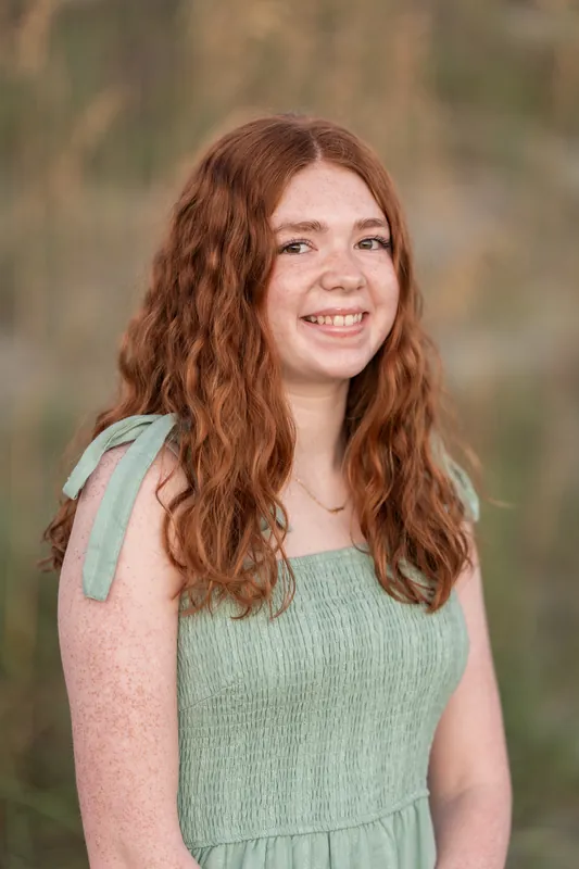 A young person with long reddish-brown waves wears a sage green sleeveless dress and smiles against a soft, blurred background.