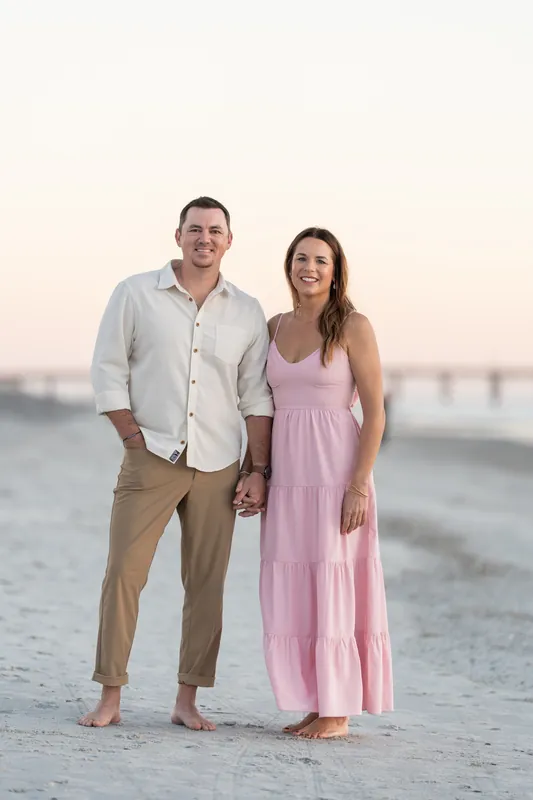 Kelly and Jereme stand barefoot on Vilano Beach at sunset, holding hands in the sand with a bridge visible across the water.