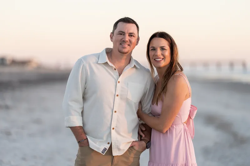 The couple smiles at the camera on Vilano Beach, with one in a light button-up shirt and tan pants, the other in a pink sleeveless dress.