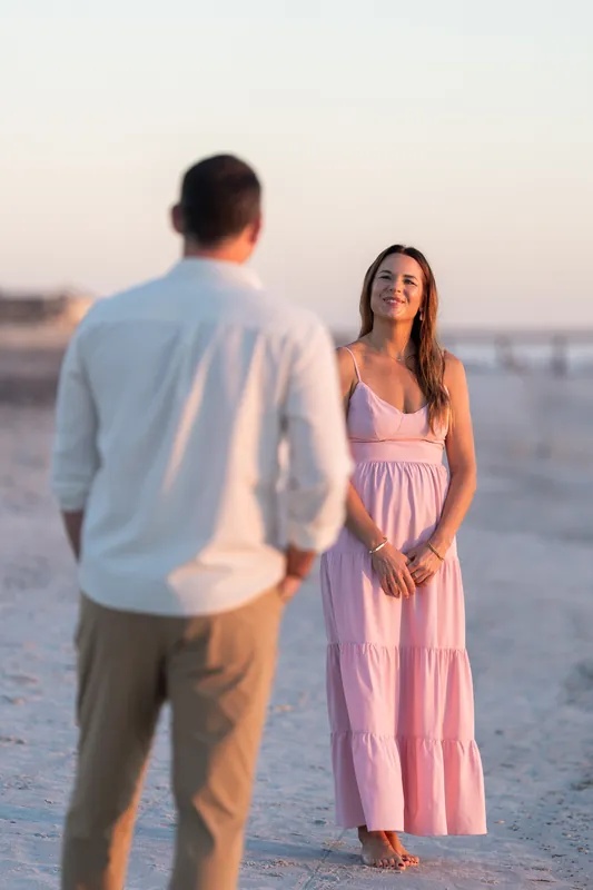Two people face each other on the beach at sunset, one wearing a light blue shirt and the other in a pink sleeveless dress.