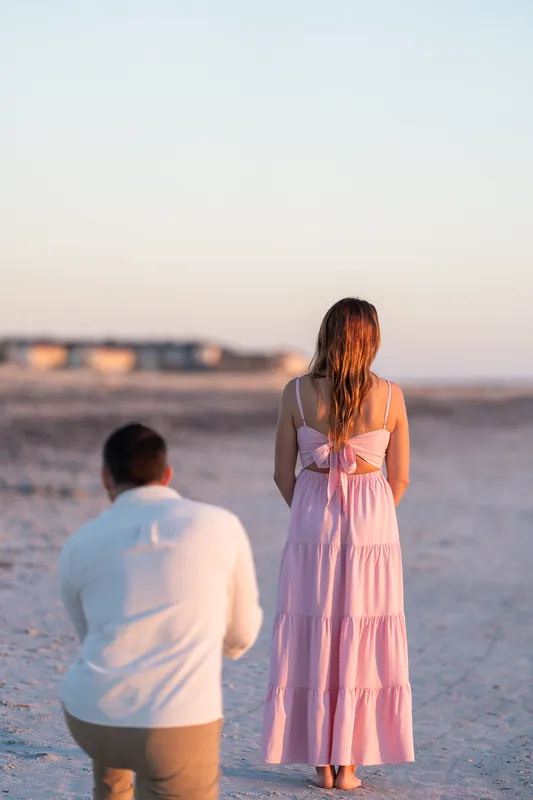 A woman in a flowing pink dress stands on the sand facing away while a man in a white shirt approaches from behind at Vilano Beach.