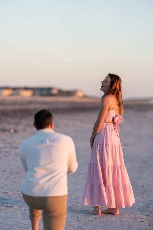 Backlit by sunset on Vilano Beach, a woman in a pink dress gazes toward the horizon as a man in a white shirt stands nearby in the sand.