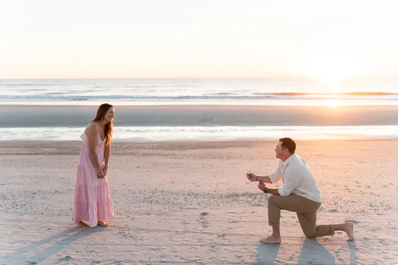 A man kneels on one knee in the sand facing a woman in a pink dress during sunset at Vilano Beach.