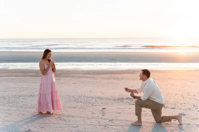 Someone in a white shirt kneels on wet sand facing a woman in a pink dress at sunset at Vilano Beach.