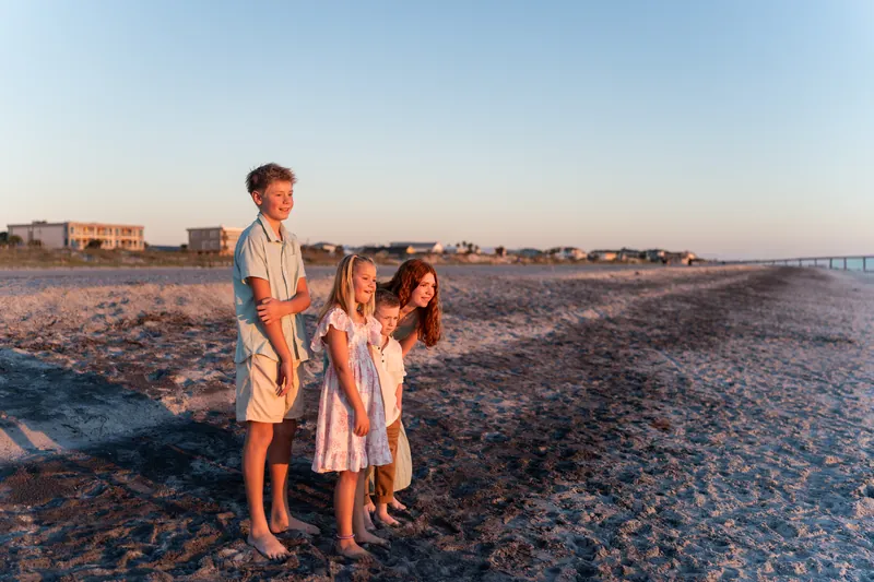 Three people stand together on the sand in soft evening light, with beach houses visible along the shoreline behind them.