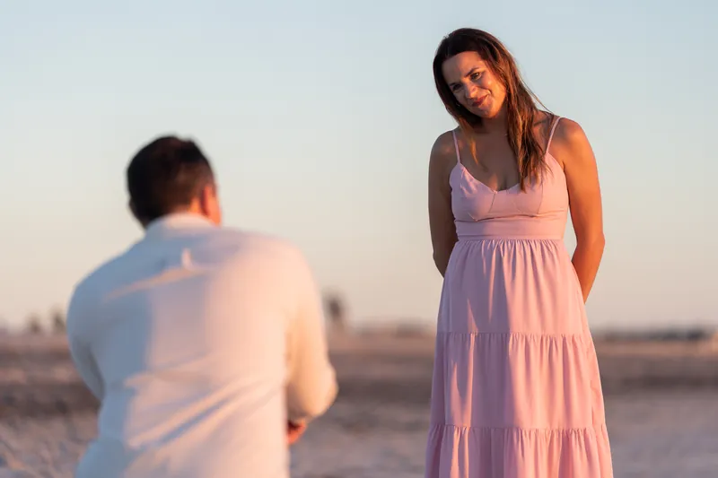 A woman in a lavender dress looks down at someone in a white shirt facing away from the camera on the beach.