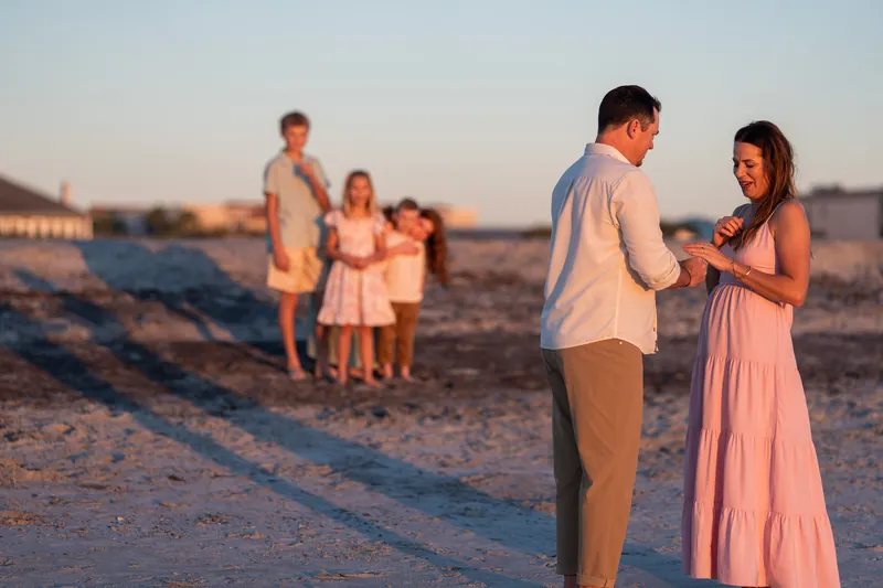 A couple holds hands while a family watches from a distance across the beach at dusk.