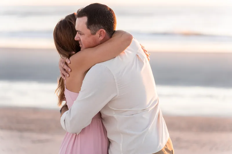 Kelly and Jereme embrace at sunset along Vilano Beach, with soft waves visible behind them.