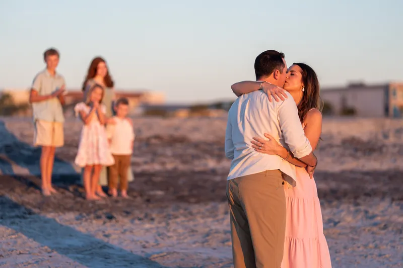 Two adults kiss while four children stand watching on the sand at Vilano Beach during golden hour.