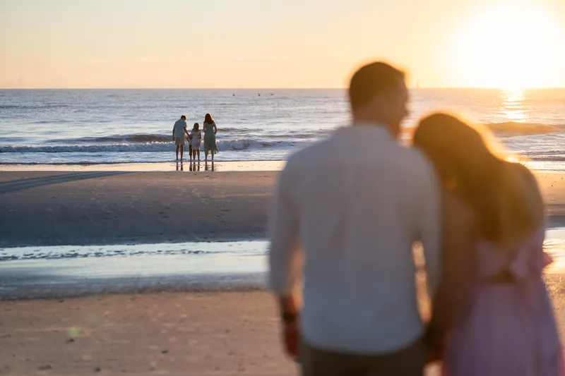 Two adults stand in the foreground watching three people pose together in the shallow water at sunrise.