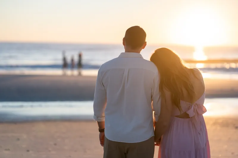 Kelly and Jereme face the ocean with their arms around each other as the sun rises behind them.
