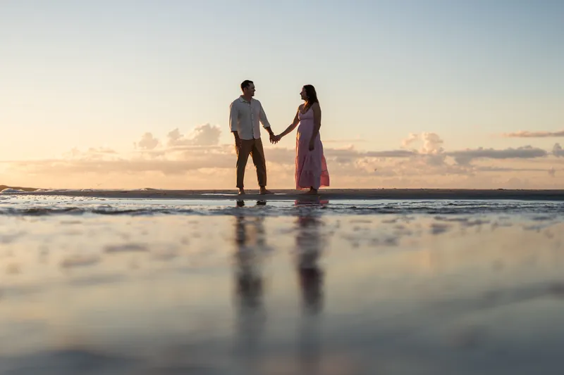 Two people hold hands in the shallow surf at Vilano Beach with the sunrise reflecting on the wet sand.