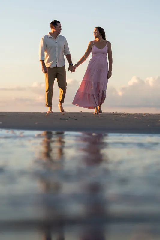 Kelly and Jereme walk hand in hand along the wet sand at Vilano Beach at sunset, ocean waves behind them.