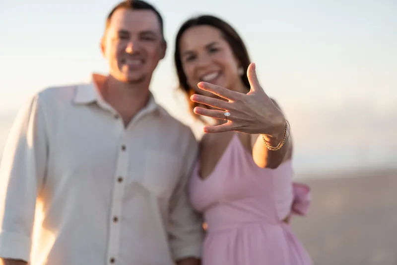 Kelly and Jereme smile at the camera, displaying a ring on an outstretched hand, golden hour light across their faces.