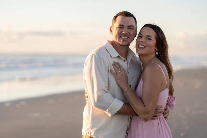 Two people embrace on the beach at Vilano Beach, both laughing, with calm ocean waves and soft sky behind them.