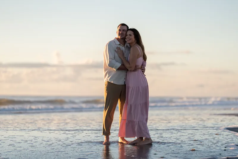 A couple stands together in shallow water at Vilano Beach, arms around each other, backlit by golden sunset light.