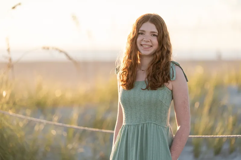 Person with reddish-brown hair in a sage green sleeveless dress smiles at Vilano Beach at golden hour, with marsh grasses behind her.