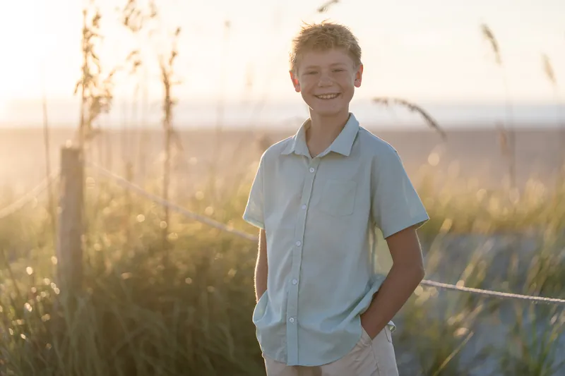 Child in light blue button-up shirt with hands in pockets grins at the camera among tall grasses at Vilano Beach.