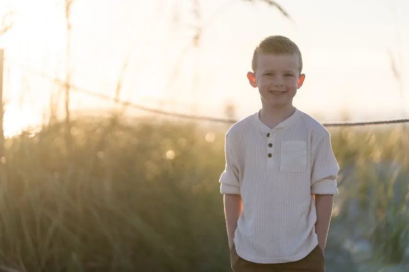Young child in white henley with rolled sleeves smiles at Vilano Beach with backlit marsh grass and rope fence behind.