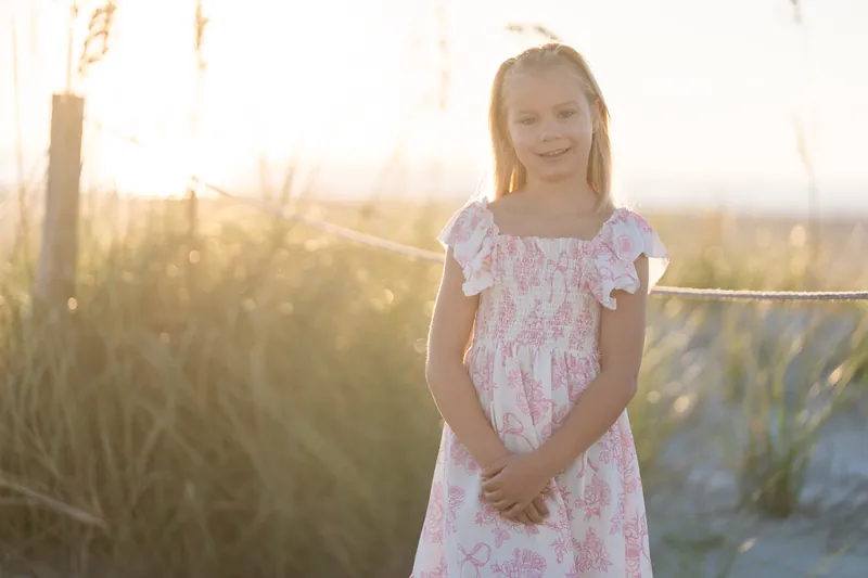Small child in lavender floral dress with clasped hands stands at Vilano Beach, glowing in late-afternoon sunlight.