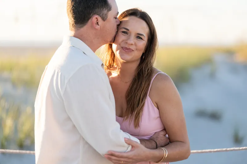 Two people kiss in profile with their hands clasped, rings visible, marsh grasses and water softly blurred behind.