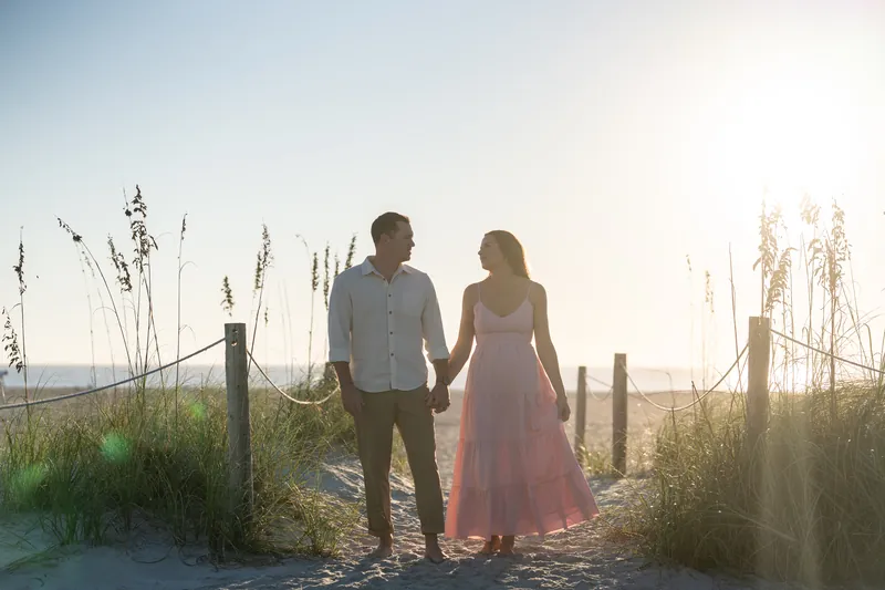 Two people walk hand-in-hand along a beach access path with sea oats and fence posts, backlit by soft evening light.