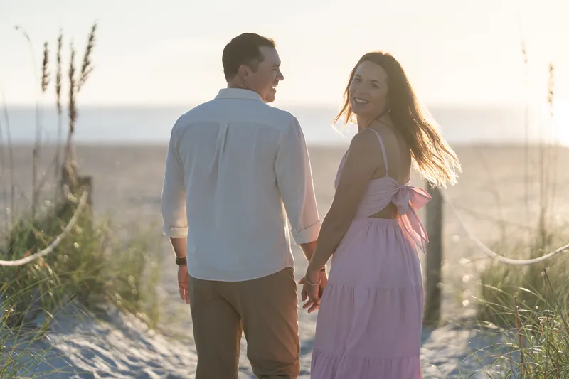 Two people walk together holding hands along the beach, the person in the light shirt looking back over their shoulder.