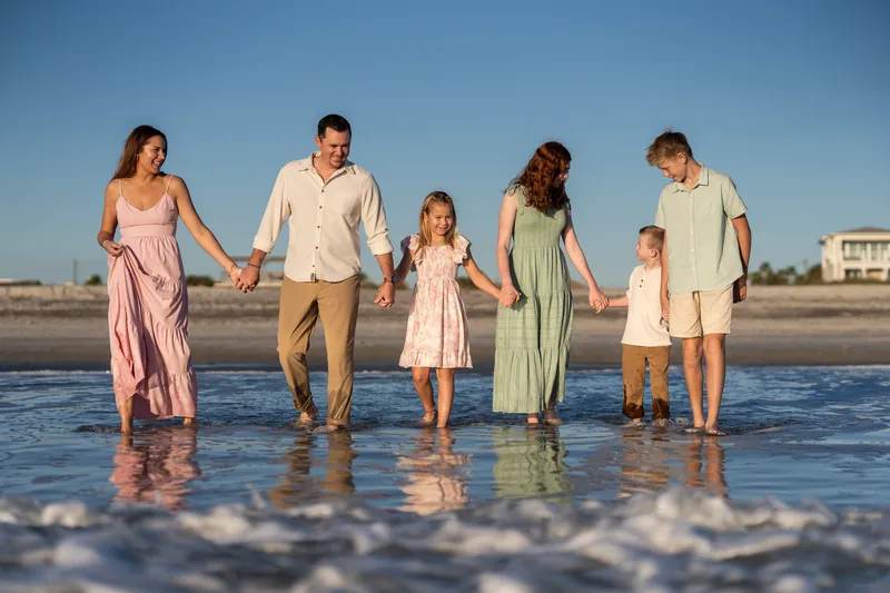 Six family members wade into shallow ocean waves at Vilano Beach in coordinated pastel outfits on a clear sunny day.
