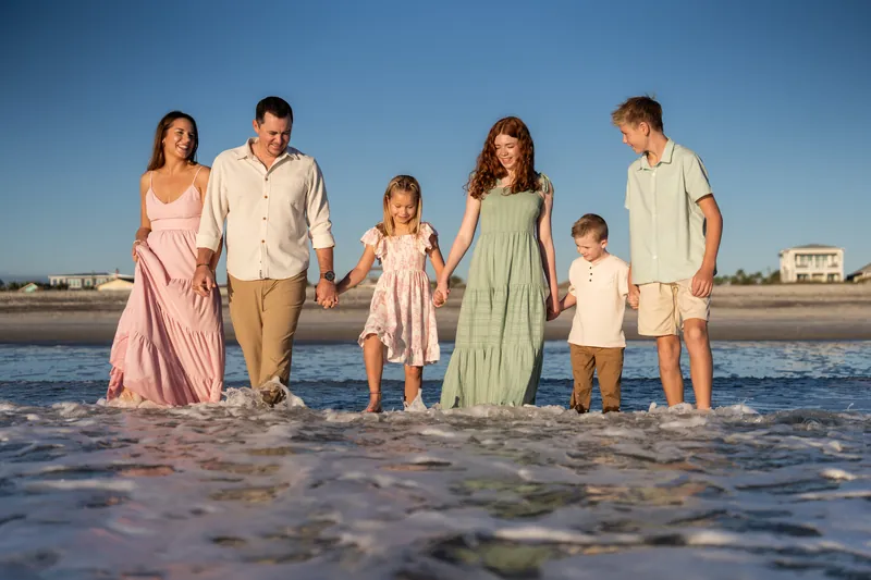 Family of six stands together in ankle-deep water at Vilano Beach, dressed in soft neutral and pastel tones under blue sky.