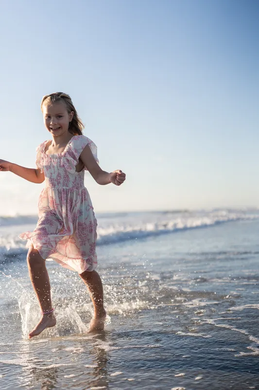 A child in a patterned dress runs through shallow water at Vilano Beach, arms outstretched.
