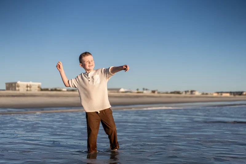 A child stands in ankle-deep water at Vilano Beach with arms spread wide, facing the camera.