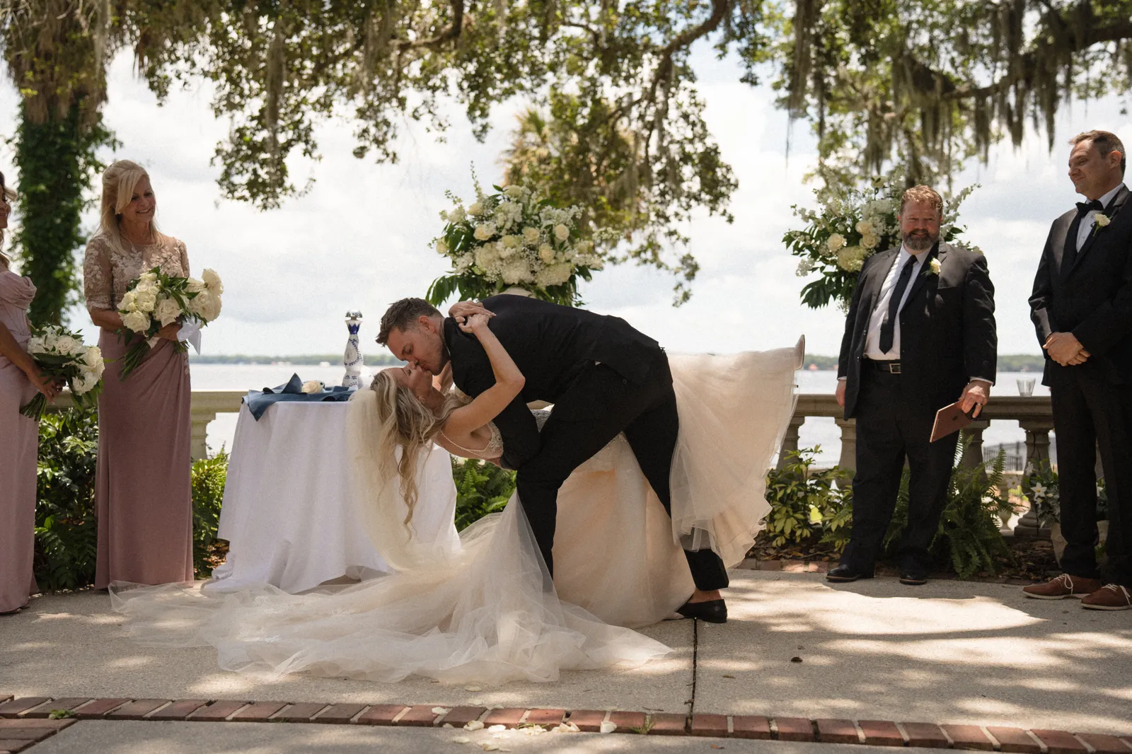 Groom dips bride during outdoor waterfront wedding ceremony under tree