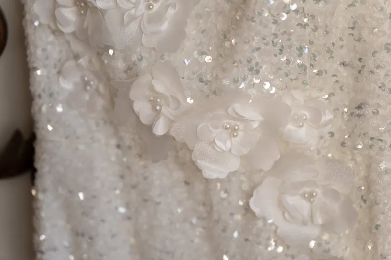 Close-up of white sequins and three-dimensional floral appliqués on shimmering wedding dress fabric.