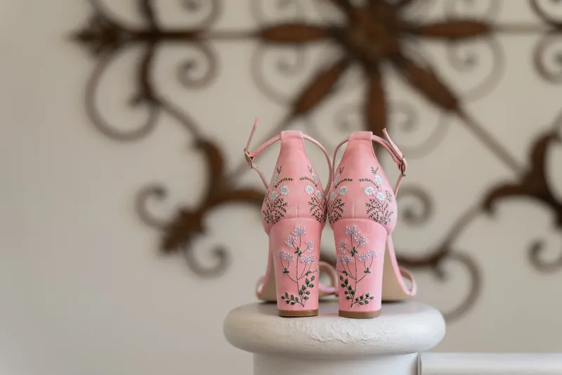 Pink embroidered heeled shoes with floral details sit on white pedestal against decorative metalwork backdrop.