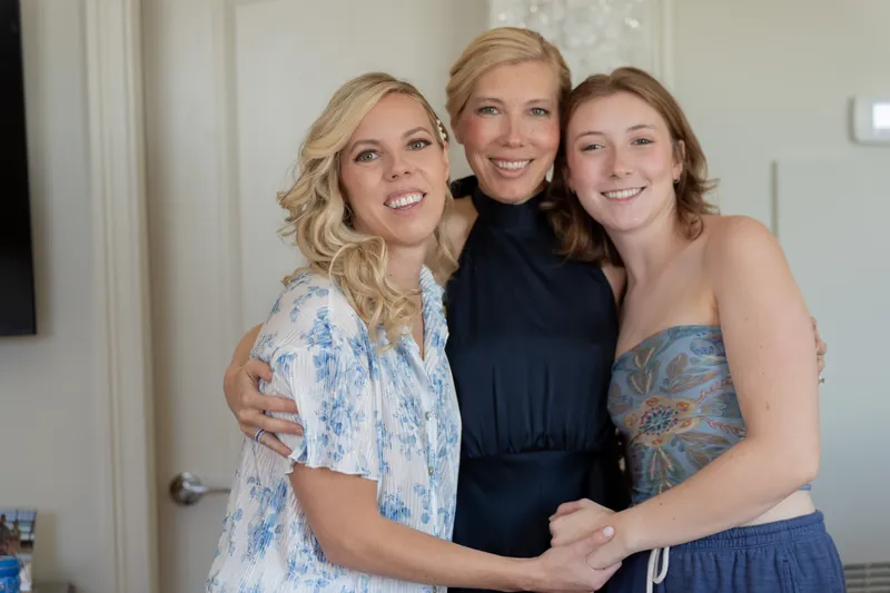 Three women smile together indoors: one in blue-and-white patterned shirt, one in black top, one in blue dress.