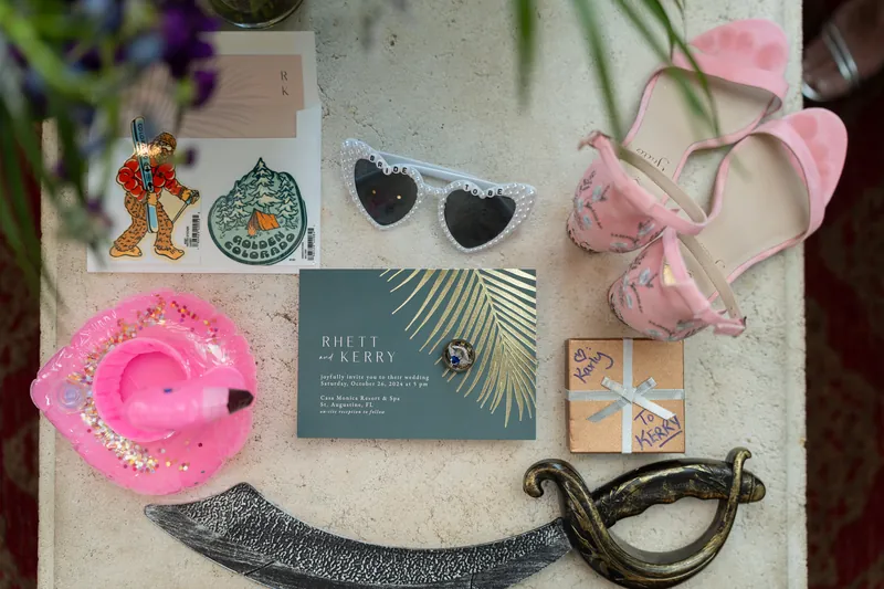 Wedding day flat lay includes pink velvet clutch, stationery, heart-shaped sunglasses, pink carnations, and decorative