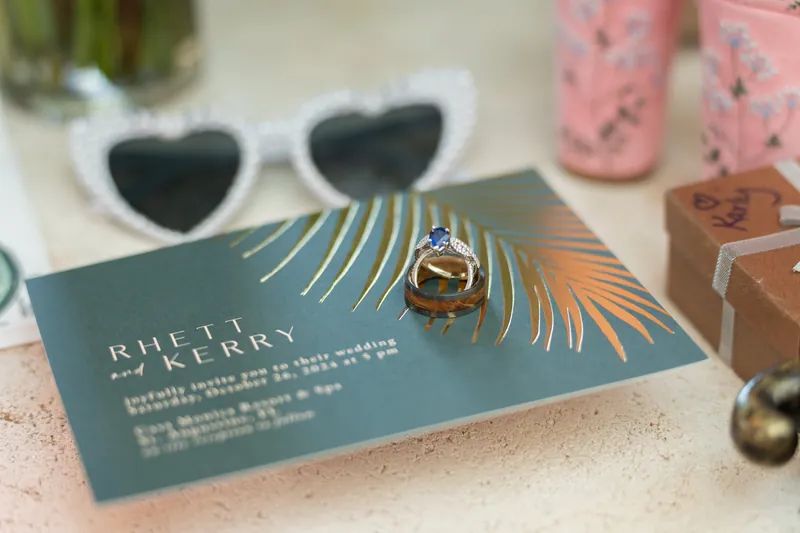Heart-shaped white sunglasses sit next to a teal wedding card with gold rings displayed on a palm leaf detail.