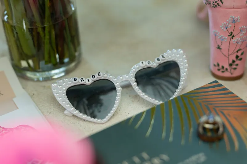White heart-shaped sunglasses spell "LOVE" as they rest beside a teal card and gold rings on a decorative surface.