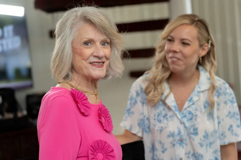 Two women in a cream-colored interior smile at each other—one wearing bright pink with ruffled sleeves, the other in a blue