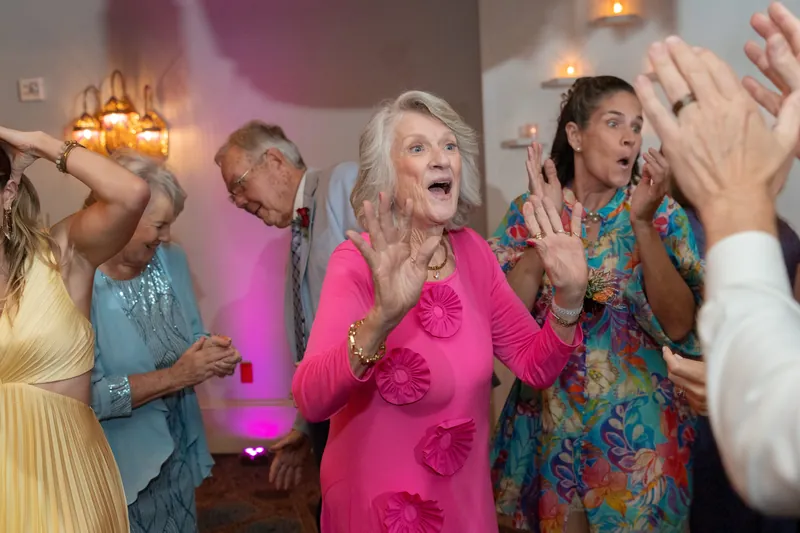 A woman in a bright pink dress laughs and dances surrounded by wedding guests in the ballroom.