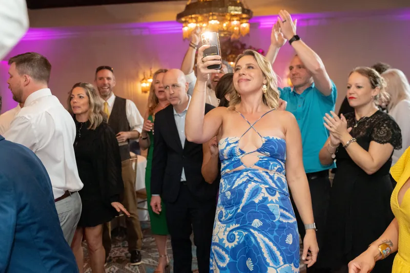 A woman in a blue patterned dress raises a chandelier overhead while dancing with guests around her.