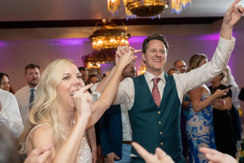Rhett in a navy vest and tie raises his arm while dancing next to a woman in a white sequined dress.