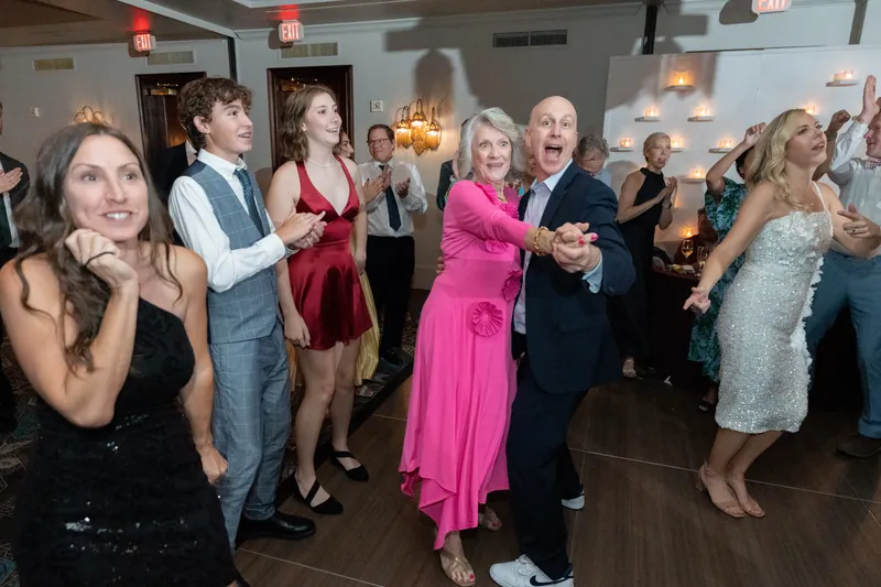 Multiple guests dance on the ballroom floor, including a woman in pink and others in various formal attire.