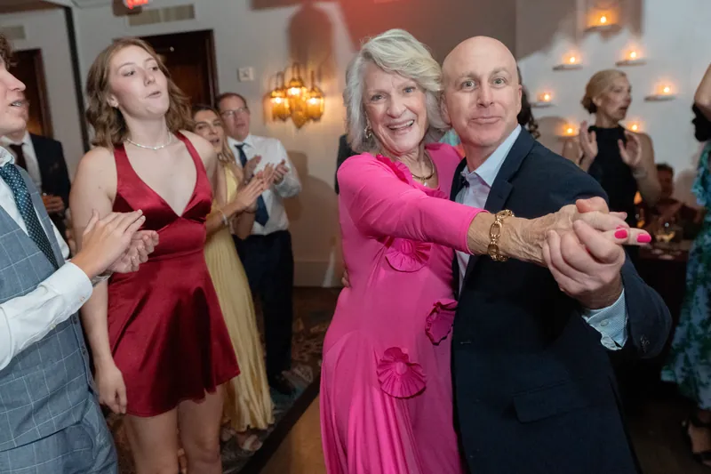 An older woman in a pink dress dances with a man in a dark suit as other guests watch and celebrate.